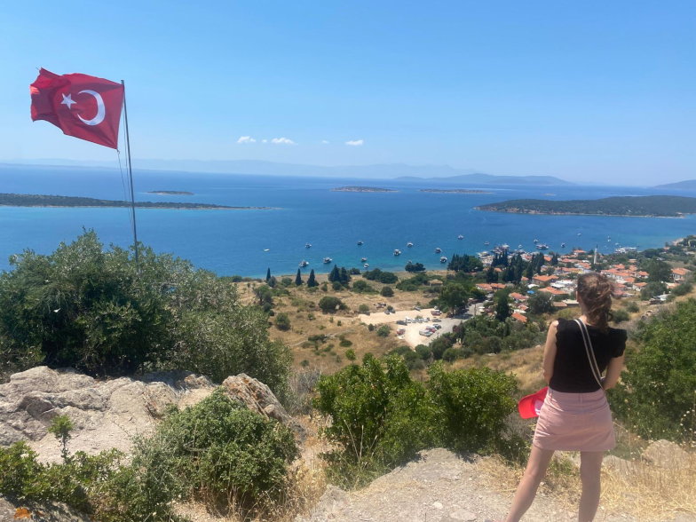 Frau blickt vom Aussichtspunkt der antiken Stadt Erythrai auf die Bucht, im Vordergrund eine türkische Flagge